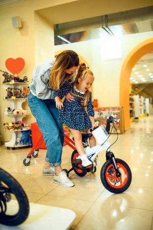 Mom and pretty little girl buying bicycle in kids store. Mother and adorable daughter near the showcase in toyshop, happy childhood, family makes a purchase in shop