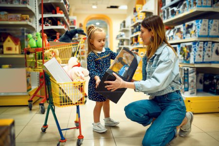 Mother and pretty little baby buying toys in store. Mom and adorable daughter near the showcase in toyshop, happy childhood, family makes a purchase in shop