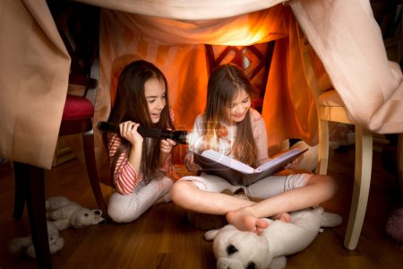 cute girls sitting under blankets and reading book with flashlig