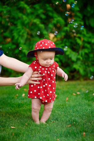 Cute funny happy baby in a red dress making his first steps on a green grass in a sunny summer garden