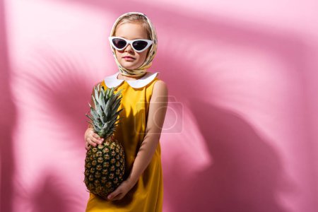 trendy girl in headscarf and sunglasses holding pineapple on pink 