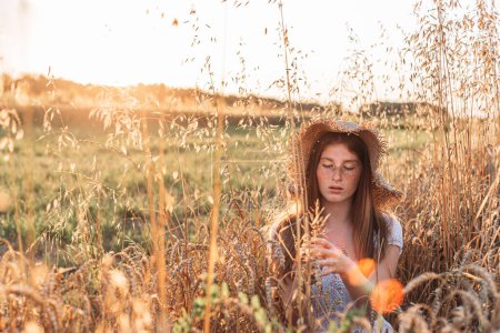 Portrait of young, teen, ginger girl with freckles in the wheat field wearing a summer hat.