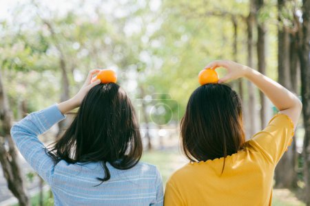 两个女孩站在一起，把橙子放在公园的头上.