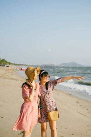两个女孩站在海滩上，戴着太阳镜的女孩指着海里漂亮的小船.