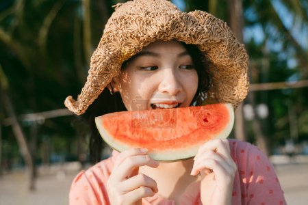 一个戴着褐色帽子的女孩站在海滩上吃西瓜.