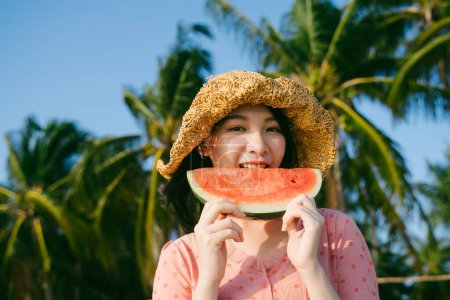 一个戴着褐色帽子的女孩站在海滩上吃西瓜.