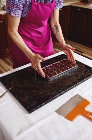 粉红围裙上的搅拌机拿着装满液体加热巧克力块的巧克力模子，放在大理石表面上。制作豪华巧克力庆祝世界巧克力日
