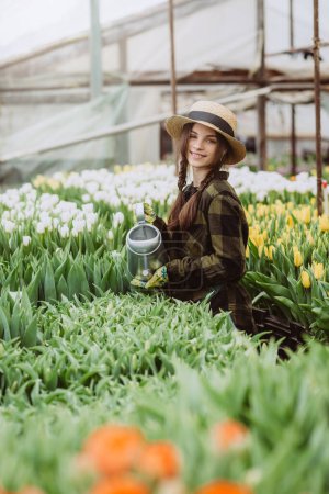 A woman gardener in a hat and gloves waters a flower bed of tulips using a watering can. Gardening hobby concept. Soft selective focus, defocus. Artistic noise.