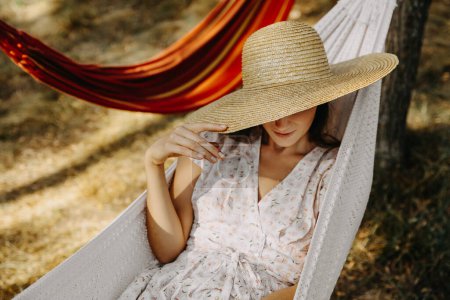 portrait of beautiful woman in dress and straw hat relaxing in hammock in forest