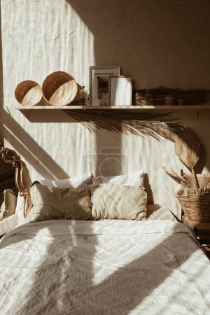 Modern boho style home interior design. Bohemian bedroom with bed, pillows, straw and rattan decorations, vintage retro mirror, palm leaf, concrete wall. Warm sunlight shadows on the wall. Hygge.