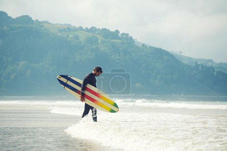 有吸引力的职业冲浪选手，抱着他走在沙滩上，英俊的冲浪者走携带他的冲浪板与背景，海洋的冲浪板过滤图像