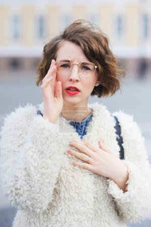 年轻漂亮的短视的女孩穿着白色夹克盯着通过他的眼镜在城市街头，特写